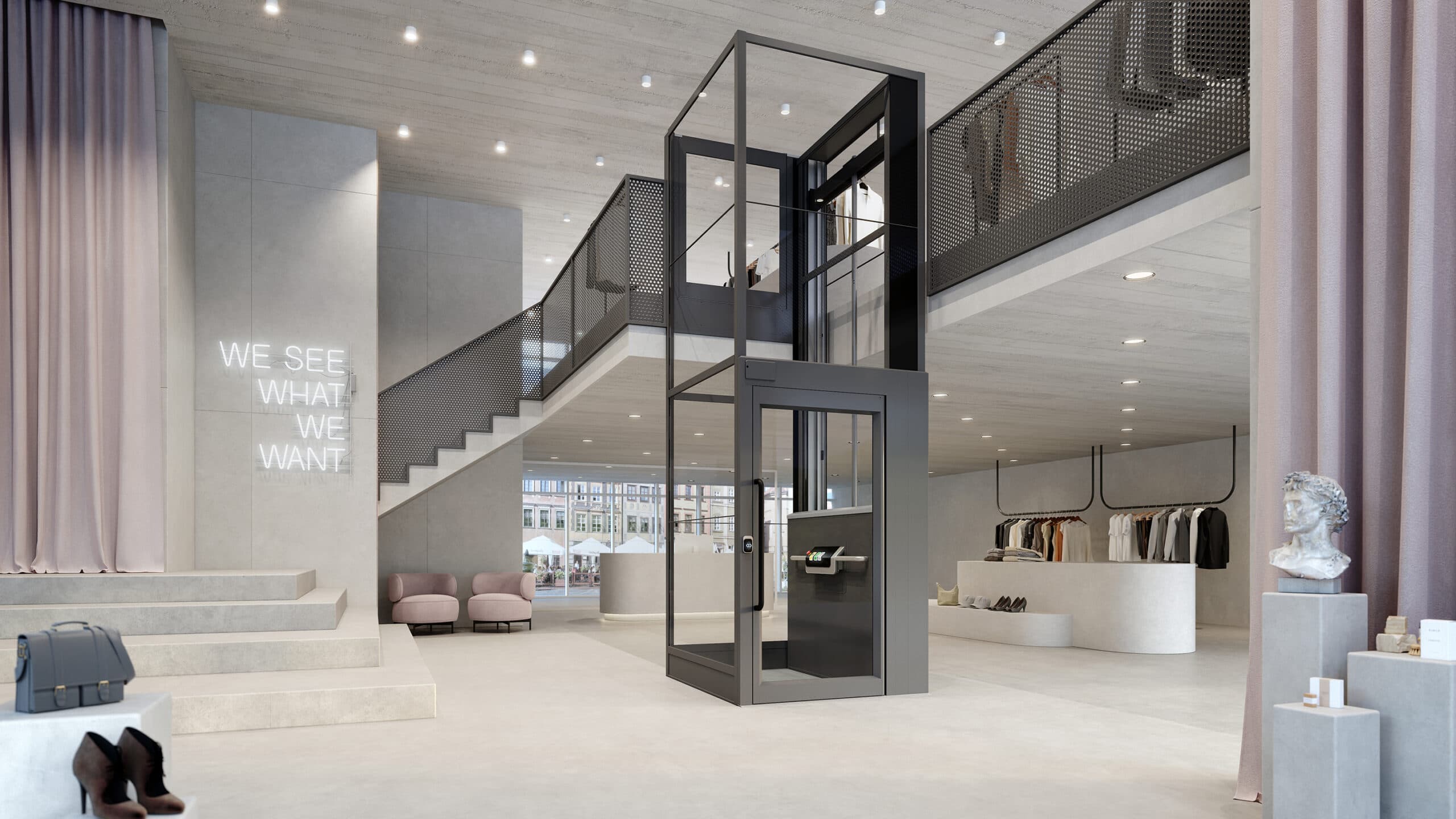 Grey Kalea Luna platform lift with a glass shaft and a gate on the top floor, installed against a mezzanine floor in a spacious clothing store with concrete walls, dusty pink textile accents and furniture and a white neon sign saying "We see what we want"