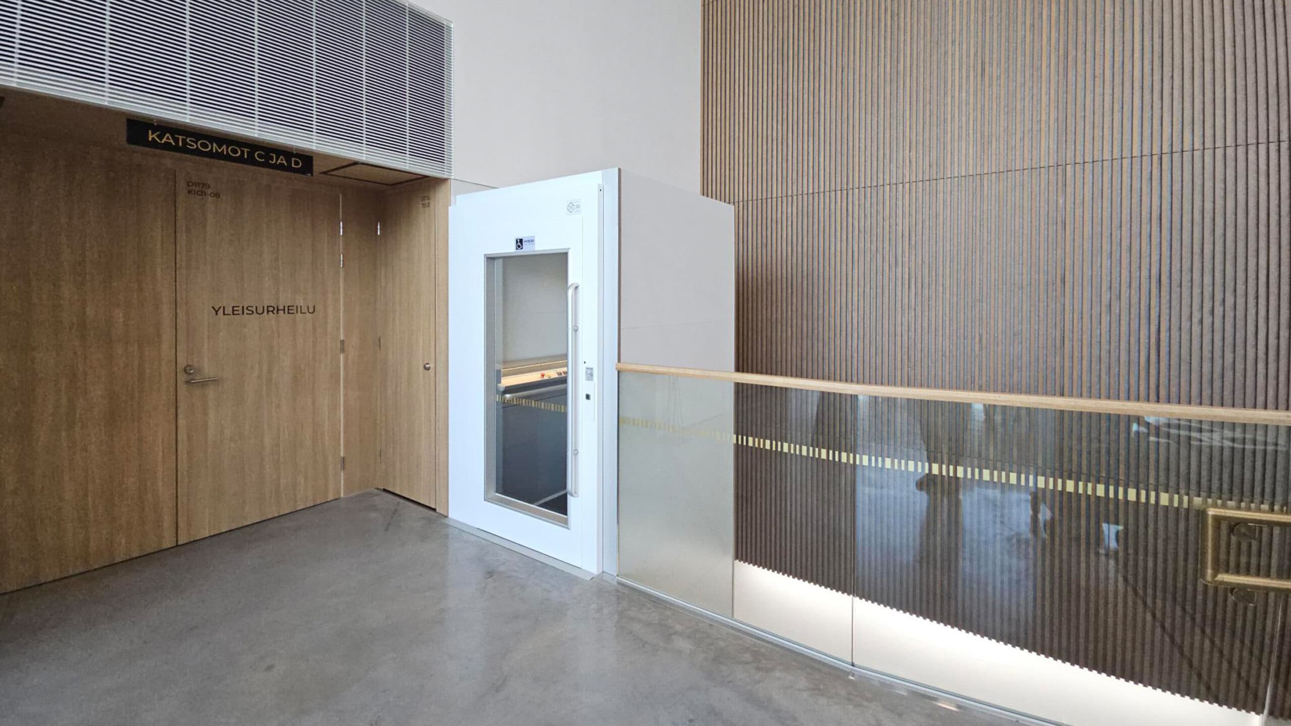 White Kalea A4 Primo wheelchair platform lift with white steel shaft seen from the top floor of a big, public hall with glass railings, wood panel walls and grey concrete floors.
